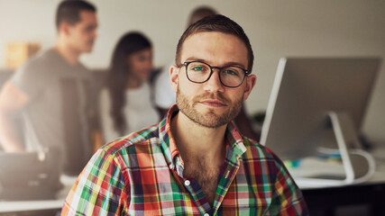 Young businessman wearing glasses, seriously looking directly into the camera, while his three diverse colleagues are in the background talking - obrazy, fototapety, plakaty