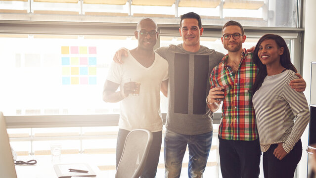 Four young diverse entrepreneurs stand side by side in an office community. They look into the camera smiling while they have their arms around each other's shoulders