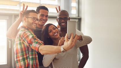 Four young diverse entrepreneurs stand in an office, and take a silly selfie together. They laugh...