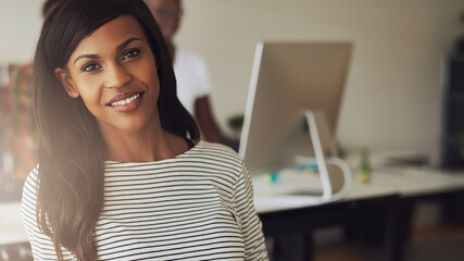 A young black female entrepreneur sits in an office and looks focused into the camera while smiling. Her diverse colleagues stand behind her and look at a computer screen