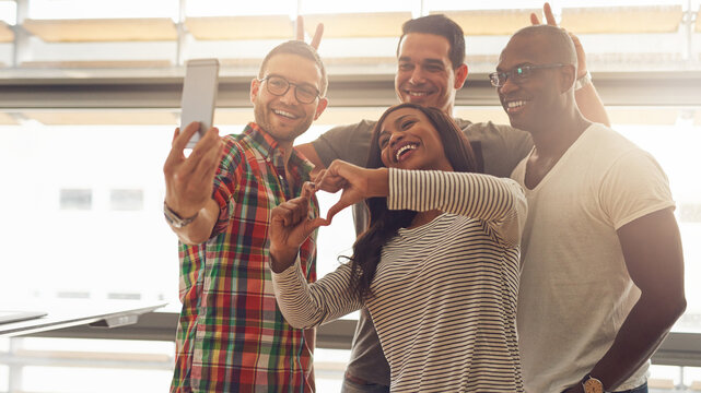 Four young diverse entrepreneurs stand in an office, and take a silly selfie together. They laugh and make funny gestures with their hands