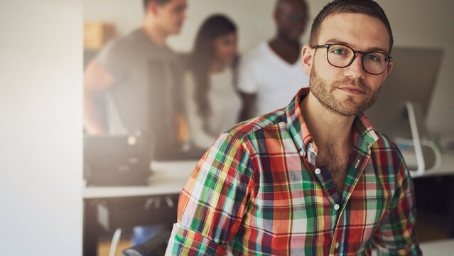 A young entrepreneur man sits in an office and looks focused into the camera, while his diverse colleagues stand behind him and look at a computer screen