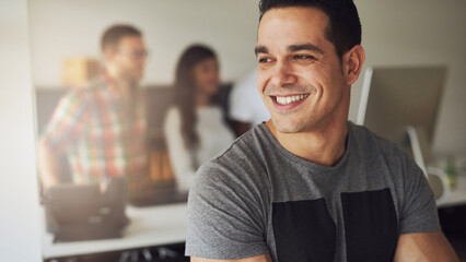 Happy athletic man sits with his arms crossed, smiling while his diverse colleagues stand in the background and look at a computer screen.