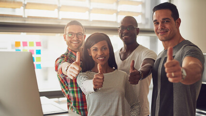 A group of diverse casually dressed business people stand together and look smiling directly into the camera while giving thumbs up. 