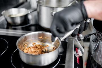 Close-up of a person in black gloves cooking food in a stainless steel pan on a modern stove. The focus is on the pan and its contents, with other pots visible in the background