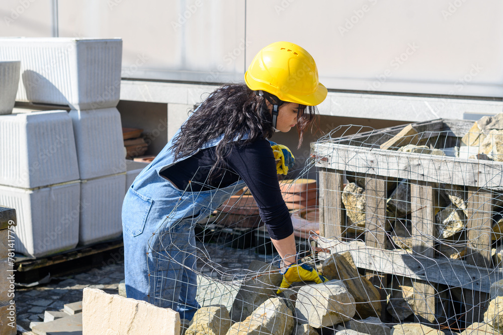 Wall mural hispanic woman work in construction site wear yellow hard hat and gloves blue jeans overall outdoors