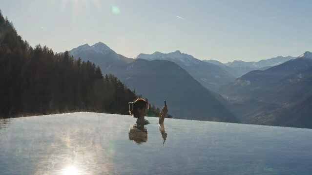 Woman takes a photo in a luxurious hot infinity pool, against a backdrop of majestic mountains. With steam rising and serene waters, she captures perfect blend of relaxation and breathtaking scenery.