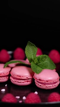 Pink sugar granules falling on rotating fresh macaron cookies. Traditional french desserts from almond dough. Pink macaron cakes, raspberry fruits and green mint leaves against black background.