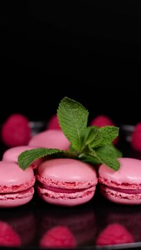 Fresh macaron cookies, raspberry fruits and green mint leaves on a plate. Traditional french desserts from almond dough. Pink macaron cakes with raspberry flavour rotating against black background.