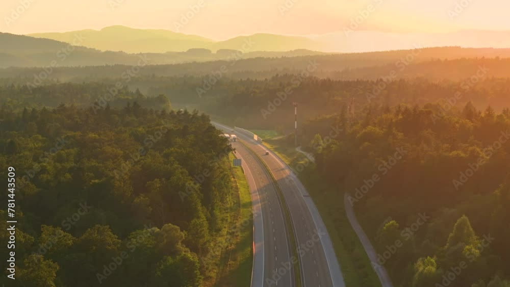 Wall mural AERIAL, LENS FLARE: Morning light spills over hilly wooded landscape and highway. Trucks and cars travel along the scenic motorway leading through beautiful countryside bathing in golden sunlight.