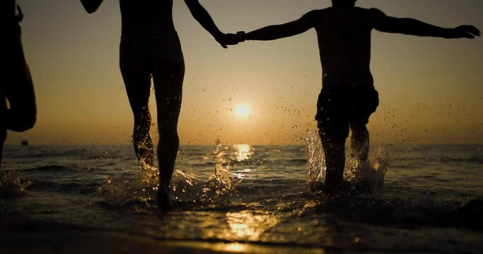 Silhouette of three friends holding hands and running in the ocean waves against a serene sunset. Splashing sea water. Recorded at 120fps on cinema camera.