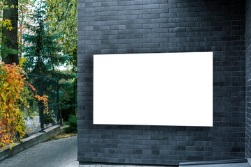 Black brick wall with blank white billboard for advertising in outdoor setting. Autumn foliage adds touch of color to modern urban landscape - obrazy, fototapety, plakaty