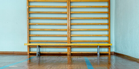 Wooden gym wall bars and bench set against pale blue wall in clean sports hall. Polished parquet flooring reflects simple functional design of workout space