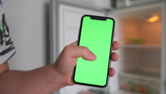 Close-up hand marking shopping list on phone with green screen chromakey against background of an open empty refrigerator. Ordering food. 
