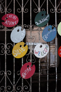 Vertical photo. Handwritten Welcome sign in English, Georgian, Italian, Spanish, French on oval wooden board hangs on old metal fence with peeling white paint. Artist's palette. Concept of entry, sale