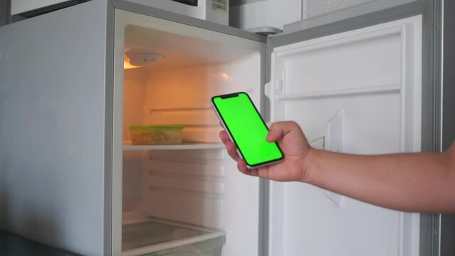 Smartphone with green screen in hand of caucasian man makes purchases in an online store on blurred background of an open empty refrigerator. Copyspase.