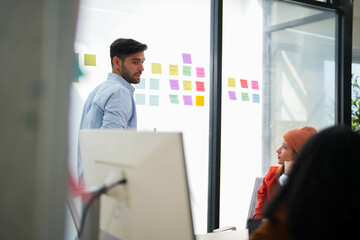 Confident male leader conducts a workshop, explaining ideas with sticky notes on a glass wall to colleagues.