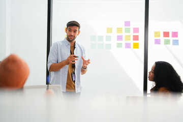 Confident male leader conducts a workshop, explaining ideas with sticky notes on a glass wall to...
