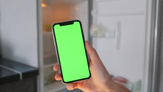 Fingers of young caucasian woman scroll green chromakey screen of smartphone against background of an open refrigerator with empty shelves, using food delivery applications.