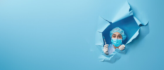 Concentrated female nurse holding a syringe peeks through a blue paper rupture