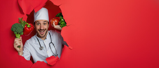 Charismatic male chef with fresh greens and tomato smiling through a torn hole in a red paper background, stethoscope visible