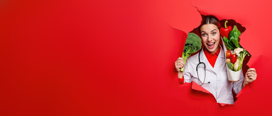 Happy female chef with a stethoscope bursts through a red background, holding a variety of greens and vegetables