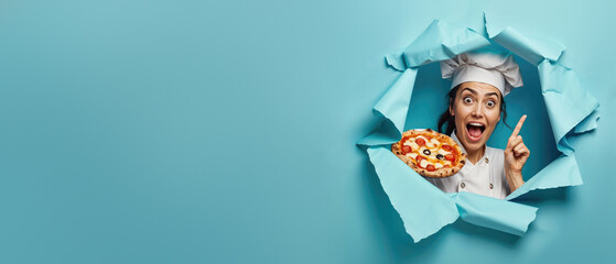 A cheerful chef making a point gesture with a pizza in hand, emerges from a torn blue background