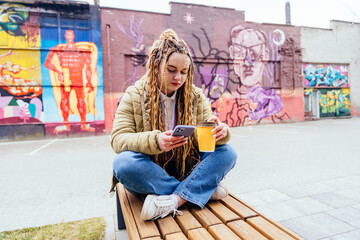 Obraz na płótnie Canvas Thoughtful sad young hipster girl using smart phone against graffiti wall