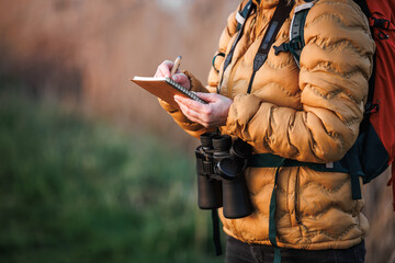 Woman ornithologist with binoculars writes details of bird behavior, migration and nesting sites in nature in a notebook. Bird watching outdoors