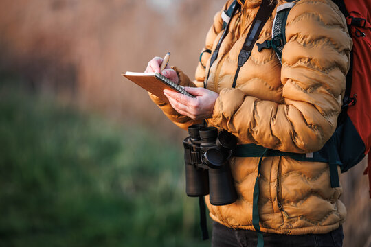 Woman ornithologist with binoculars writes details of bird behavior, migration and nesting sites in nature in a notebook. Bird watching outdoors