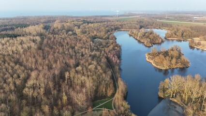 Aerial view of a serene lake surrounded by autumnal forest with walking paths and distant wind turbines on the horizon, showcasing a blend of natural beauty and renewable energy.