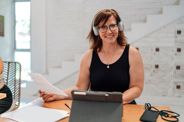 Smiling professional Caucasian woman with headphones in a modern office, reviewing documents, illustrating the merger of traditional business practices with contemporary technology