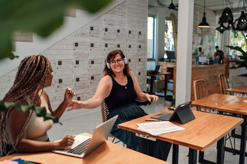 joyful interaction between two businesswomen at office table, one African-American with laptop, other Caucasian wearing headphones, in well-lit workspace, teamwork and positive corporate culture