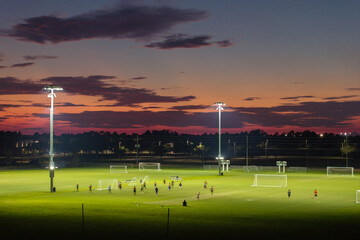 Illuminated public sports arena in North Port, Florida with people playing soccer game on grass football stadium at sunset. Outdoor activities concept - obrazy, fototapety, plakaty