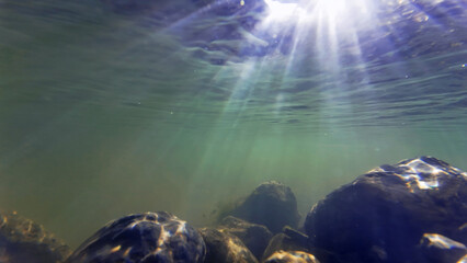 Unterwasseraufnahme vom Grund eines Flusses in dem die Lichtstrahlen der Sonne in Wasser fallen