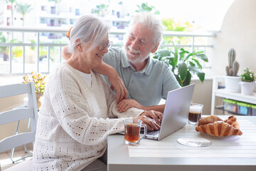 Smiling bonding senior retired couple browsing with laptop while enjoy breakfast together sitting...