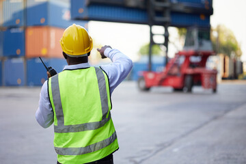back view African worker or engineer using walkie talkie and showing gesture to crane car in containers warehouse storage
