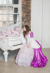 Young beautiful sad woman in fantasy rococo style medieval dress sitting near piano with pink flowers