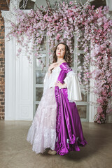 Beautiful woman in fantasy white and purple rococo style medieval dress standing near wall with pink flowers