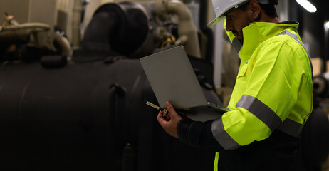 Mechanical engineer with green safety jacket holding laptop and walkie talkie working at site line of HVAC control room - obrazy, fototapety, plakaty
