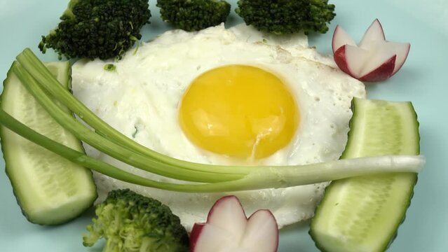 Fried egg, boiled broccoli, radishes, fresh cucumbers and green onion are on a pale blue plate, healthy breakfast