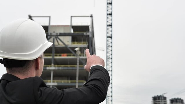 Architect pointing with hand at some details on construction, sites in the background construction crane and unfinished building Architect in business clothes and white helmet action on the street