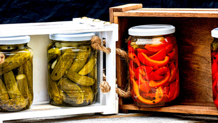 Wooden crate with glass jars with pickled red bell peppers and pickled cucumbers (pickles)...