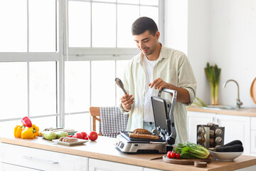 Young man cooking tasty sausages and vegetables on modern electric grill at table in kitchen