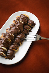 Mini pancakes with banana, poured with chocolate in a plate in bed on a dark background, top view.