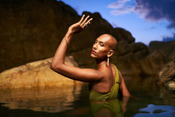 Androgynous black lgbtq fashion model dives out of water inside picturesque natural pool at night. Non-binary biethnic person poses waist deep in still water among rocks on exotic island. Pride month.