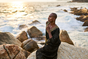Queer black person in luxury dress, jewelry sits on rocks in ocean. Lgbtq ethnic fashion model...