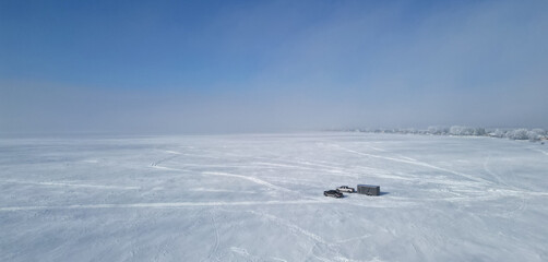 Panorama of frozen lake with traces of the off-road car. Pickup with trailer for ice fishing or hunting. Minimalist winter landscape with frosty haze.