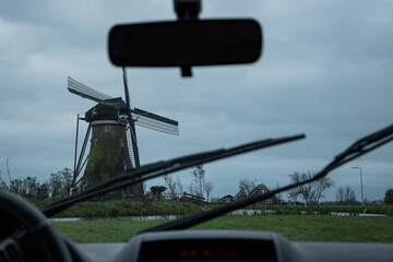 A typically miserable rainy day in the Netherlands view from inside a car. windmill and grey stormy clouds with rear view mirror, dashboard and windscreen wipers on Dutch bad weather day in Holland