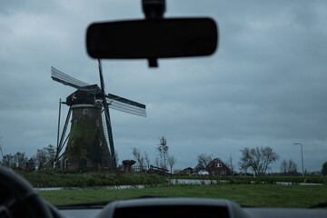A typically miserable rainy day in the Netherlands view from inside a car. windmill and grey stormy clouds with rear view mirror, dashboard and windscreen wipers on Dutch bad weather day in Holland - obrazy, fototapety, plakaty